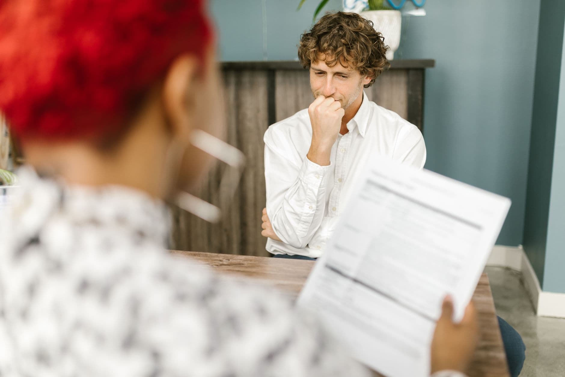 Deux personnes sont assises à une table dans un intérieur, l’une au premier plan tient des feuilles, l’autre, en chemise blanche, réfléchit.